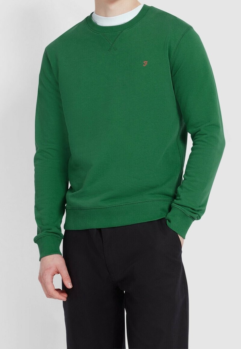 Homme portant un sweat-shirt vert à manches longues sur un maillot blanc, associé à un pantalon noir, main dans la poche.