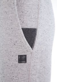 Primer plano de una tela gris moteada con un bolsillo interior de ribete negro y un pequeño parche de cuero negro en forma de cuadrado con un diseño de botón circular.