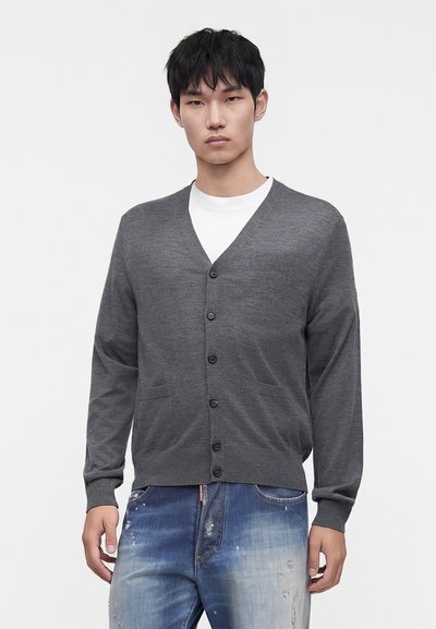 Jeune homme portant un cardigan boutonné gris foncé sur une chemise blanche et un jean bleu délavé avec un aspect usé.