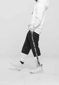 Svart och vit trackbyxor med en avsmalnande design, som har ett vitt midjeband och logoförstärkningar, ihop med vita högskurna sneakers.