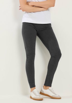 Mujer con leggings gris oscuro y zapatillas blancas, de pie con los brazos cruzados frente a una pared blanca.