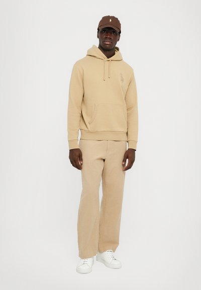 Beige hoodie med ficka fram och logodetalj, i kombination med matchande lösa byxor och vita sneakers. En brun keps fulländar outfiten.