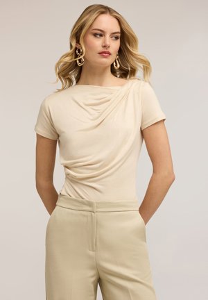 Blondynka w beżowej, drapowanej bluzce z krótkim rękawem, dopasowanych spodniach z wysokim stanem i dużych złotych kolczykach kołach, patrząca w prawo, z rękami za plecami.