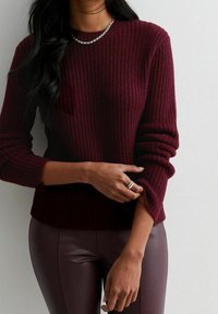 Maglione a coste color bordeaux con scollo rotondo e maniche arrotolate, abbinato a pantaloni in ecopelle marroni. Una collana in argento mette in risalto il décolleté.