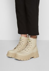 Stivaletti beige con una texture liscia, punta rotonda, suola spessa e lacci evidenti. Presentano un'etichetta con il logo sulla linguetta.