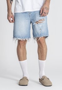 Svijetloplavi traper šorc sa rastrganim rubovima i istrošenim detaljima. Nosite s bijelim čarapama i bež sandalama bez kopčanja.