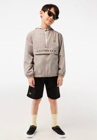 Šviesiai pilkas anorakas su juodu logotipo dryžiu, juodi šortai ir geltonai dryžuotos kojinės. Aksesuarai: juodos saulės akiniai ir juodi sportiniai bateliai.