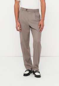 Άνδρας που φοράει ανοιχτό καφέ ραμμένο παντελόνι και μαύρα-άσπρα μοκασίνια, στέκεται μπροστά σε λευκό φόντο με τα χέρια χαλαρά στα πλευρά.