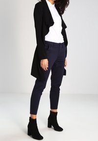 Cardigan negro de apertura frontal, camisa blanca, pantalones azul marino y botas negras de tobillo. Tejidos suaves, silueta ajustada y calzado texturizado.