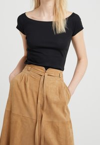 Kvinna klädd i en svart off-shoulder topp och en högmidjad beige mockaskjorta med en knytkedja, med händerna i kjolens fickor.