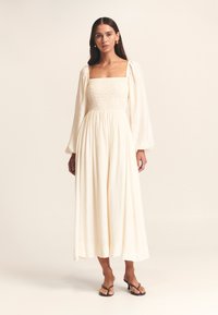 Langes cremefarbenes Kleid mit Puffärmeln, gerafftem Oberteil und fließendem Rock. Leichtes Gewebe mit weicher Textur. Schwarze Sandalen.