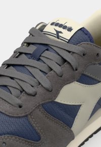 Scarpe sportive grigie e blu navy con una tomaia in camoscio e rete, caratterizzate da un accent beige prominente e lacci piatti, contrassegnate con il nome del marchio.