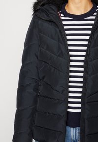 Schwarze Pufferjacke mit horizontalen Nähten, Kapuze mit Fellbesatz und Reißverschluss-Taschen, getragen über einem navy- und weiß gestreiften Pullover.