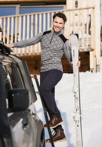 Grå mønstret langærmet top med sorte detaljer, marineblå bukser og brune snørebåndsstøvler. Holder hvide ski med bindinger, placeret på et køretøj.