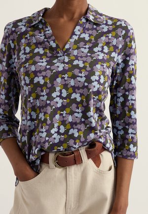 Personne portant un chemisier violet à motif floral avec des manches trois-quarts, rentré dans un pantalon beige clair avec une ceinture en cuir marron.