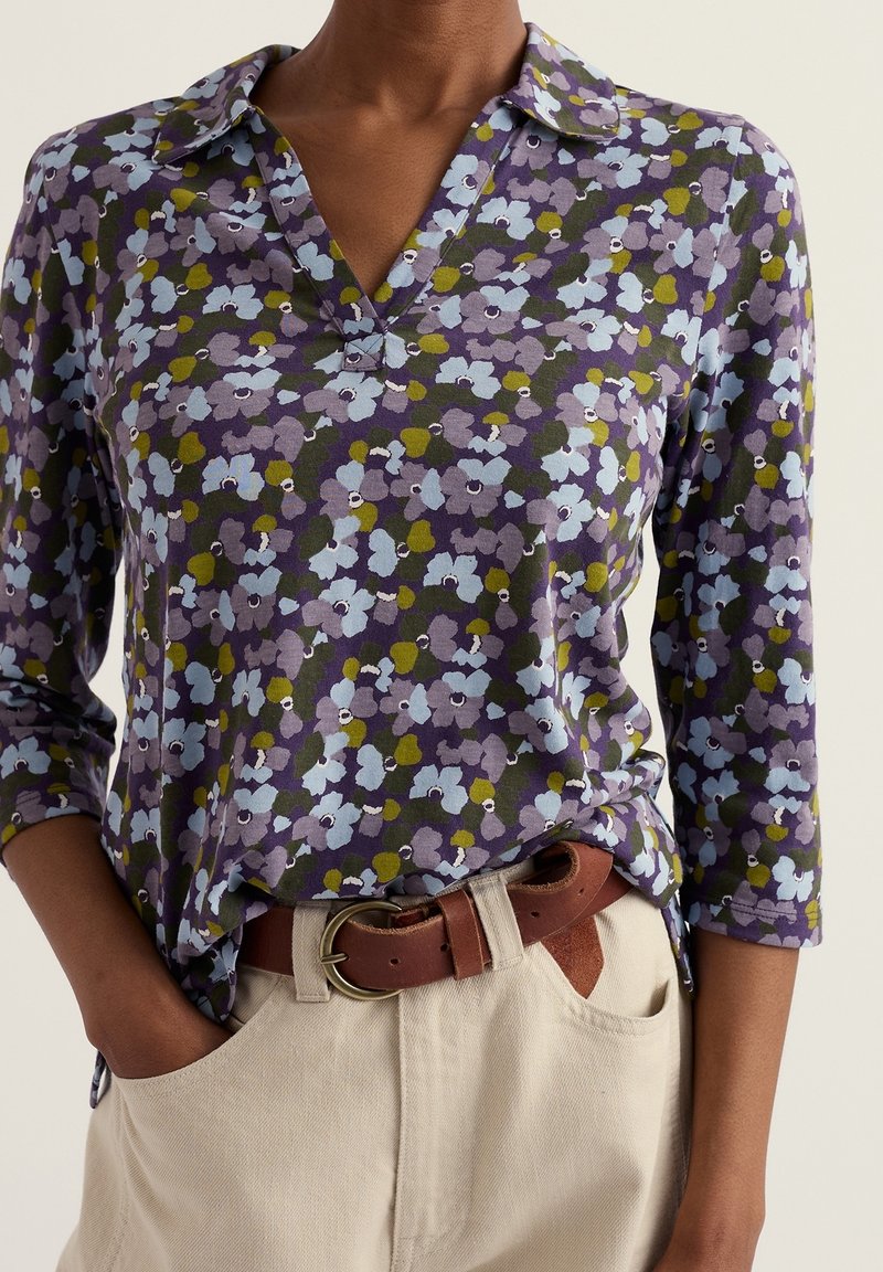 Personne portant un chemisier violet à motif floral avec des manches trois-quarts, rentré dans un pantalon beige clair avec une ceinture en cuir marron.