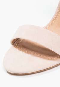 Sandale en suède beige avec une large lanière sur le pied, dotée d'un intérieur en cuir lisse et de coutures subtiles le long du bord.