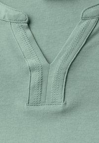 Top in cotone verde chiaro con scollatura a V e cuciture dettagliate, maniche corte e superficie liscia.