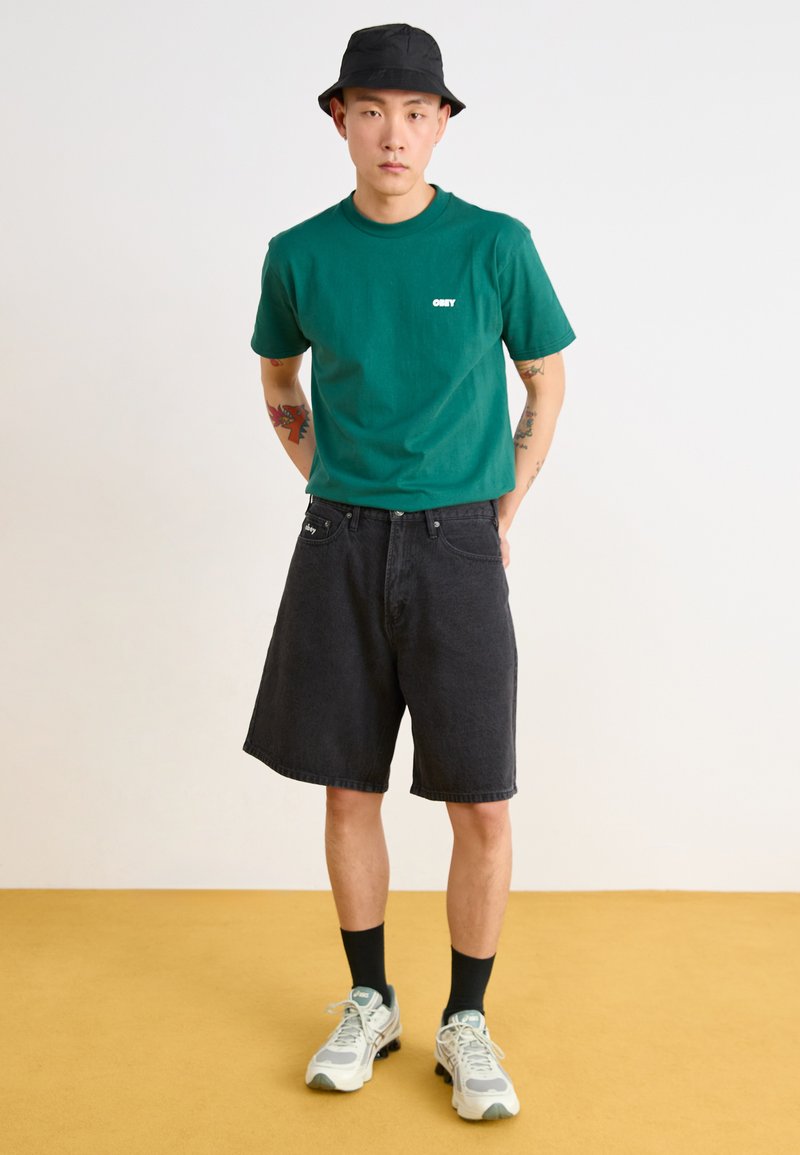 T-shirt en coton vert avec logo blanc, short en denim noir, chapeau seau noir, baskets blanches avec accents gris, chaussettes noires. Fond uni.