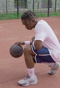 Basketballspiller iført en hvit skjorte og marineblå shorts, som croucher på banen, holder en svart basketball, grå joggesko med aksenter.