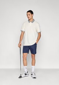 Πόλο πουκάμισο σε εκρού με γκρι γιακά και λεπτομέρειες στα μανίκια, συνδυασμένο με σκούρο μπλε σορτς. Το μοντέλο κρατάει ένα γκολφ κλαμπ, φορώντας αθλητικά λευκά αθλητικά παπούτσια.