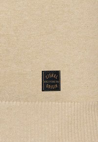 Beige gestrickter Pullover mit geripptem Saum, der ein schwarzes quadratisches Etikett mit der Aufschrift "SIGNAL" und "ORIGIN" in weißem Text trägt.