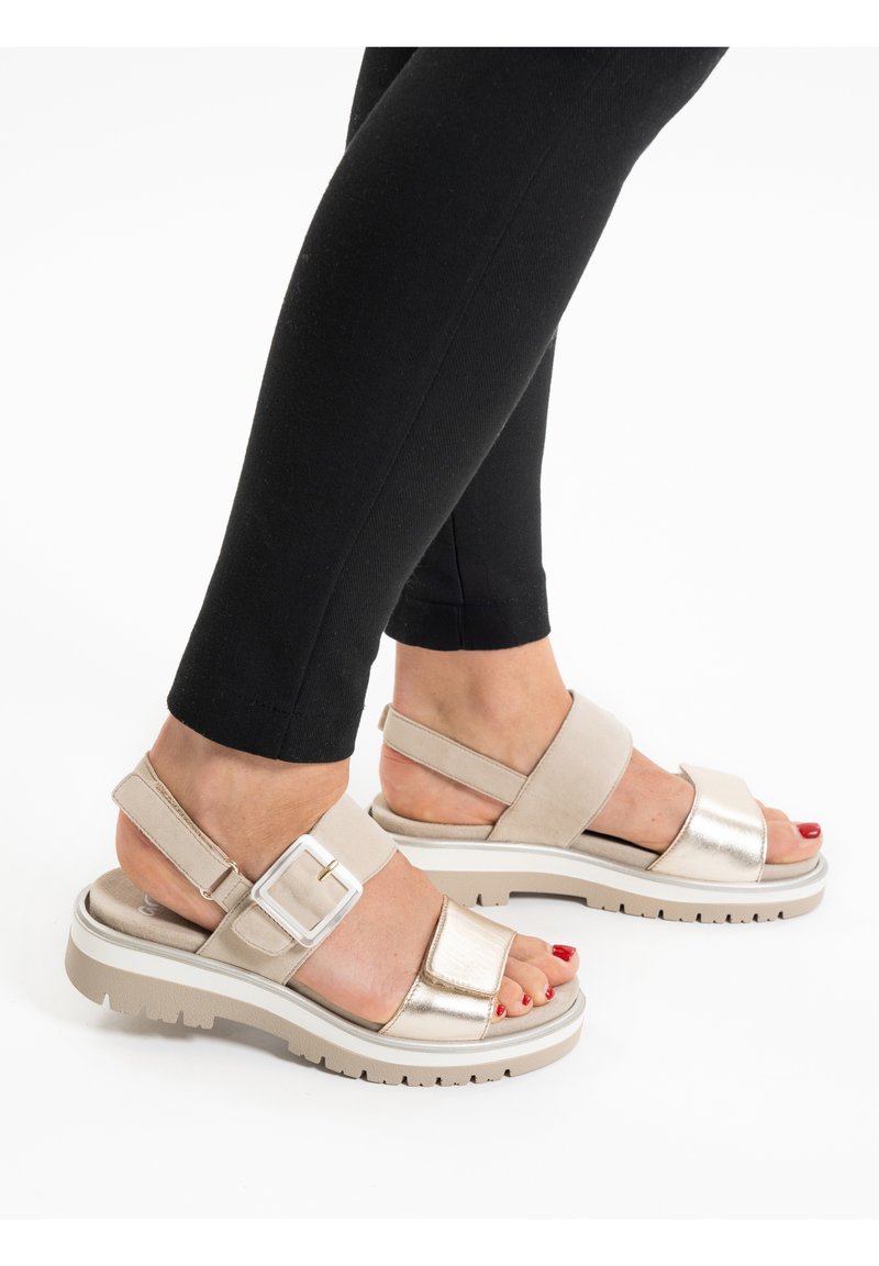 Beige sandalen met verstelbare banden, textuurmetalen accenten, een gevoerde zool en een gevormd ontwerp, gecombineerd met zwarte leggings.