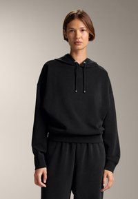 Felpa nera con cappuccio in una vestibilità comoda, con cordoncino al collo, polsini a costine e una texture liscia. Abbinata a pantaloni coordinati.