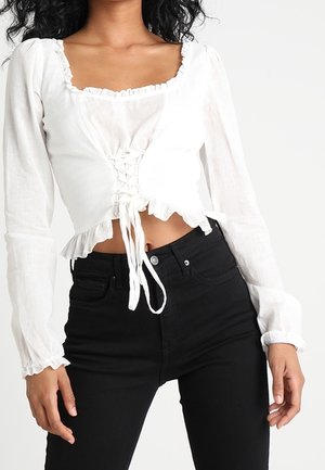Vrouw draagt een witte cropped blouse met lange mouwen en veterdetail aan de voorkant en zwarte hoge taille skinny jeans.