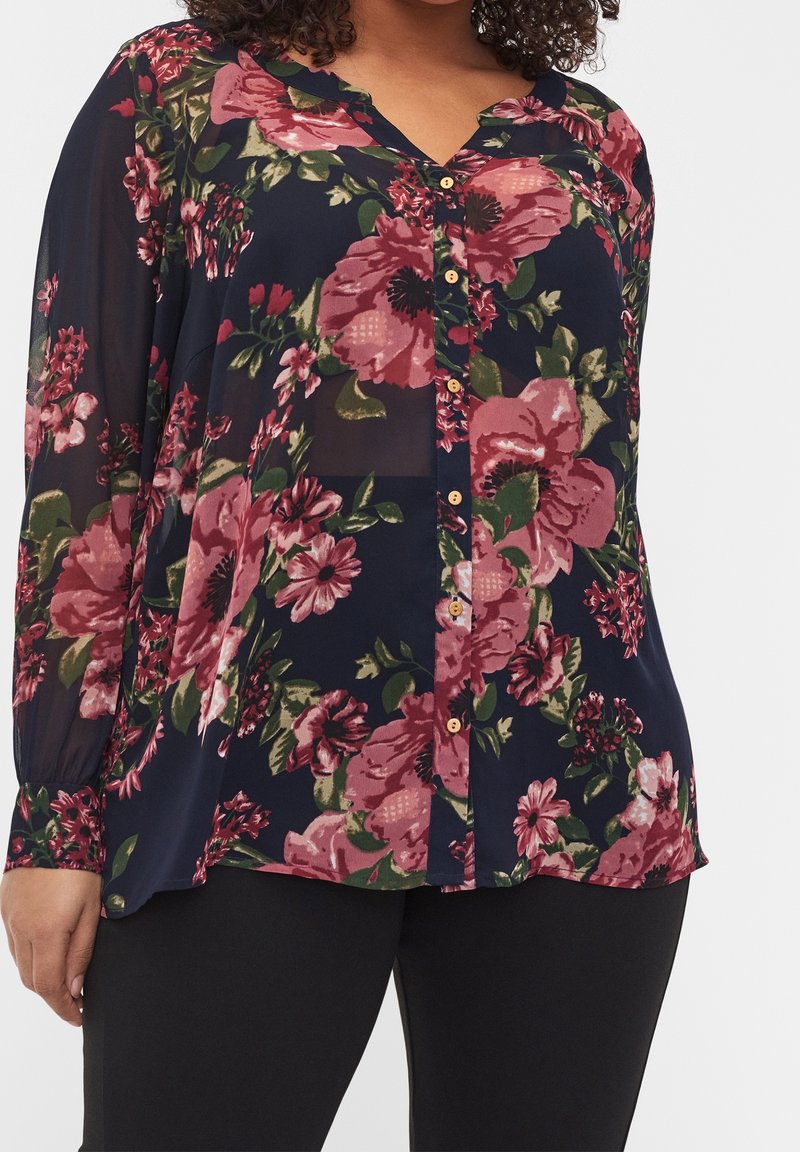 Blouse bleu marine boutonnée avec manches longues transparentes et grand imprimé floral rose, associée à un pantalon noir.