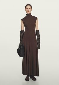 Robe marron sans manches avec un col montant, longueur midi, dotée d'une jupe fluide. Portée avec de longs gants noirs et tenant un sac à main noir.