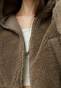 Brązowa polarowa kurtka z zamkiem błyskawicznym, z teksturowanym, puszystym wykończeniem i gładkim, jaśniejszym podszewką.