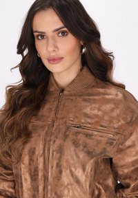 Jachetă bomber metalică de bronz cu guler ribbed, buzunar cu fermoar și suprafață texturată. Finisaj neted cu modele subtile, având un fermoar aurie.