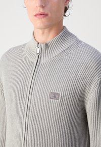 Maglione grigio a coste con zip e colletto alto in tessuto texturizzato. Presenta una piccola patch con logo sul lato sinistro vicino alla zip.