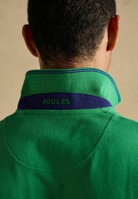Dos d’une personne aux cheveux courts et bouclés portant une chemise verte à col avec une bande bleue marine et "JOULES" brodé sur le col.