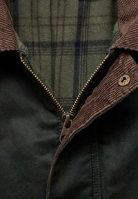 Giacca cerata scura con zip bronzo parzialmente aperta, colletto in corduroy marrone e fodera interna a quadri verdi visibile.