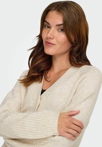 Bež pletena jopica z V-izrezom, rebrastimi manšetami in teksturirano tkanino, oblečena čez črno čipkast top. Model z dolgimi valovitimi lasmi.