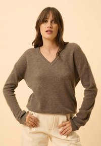 Sweter z dekoltem w serek, długim rękawem, w średnim odcieniu brązu, strukturalny splot w romby; noszony z jasnymi spodniami. Prosty design, luźny krój.