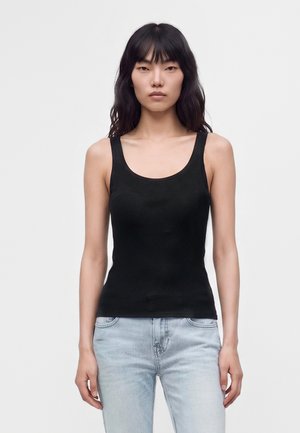 Femme aux longs cheveux noirs portant un haut noir sans manches et un jean bleu clair, debout face à l’avant sur un fond uni.