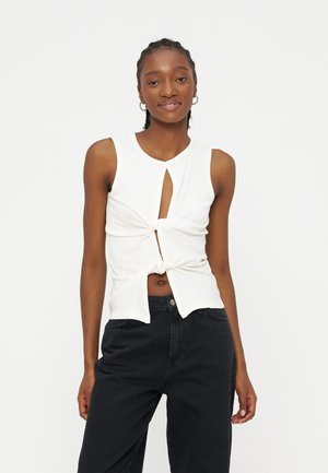 Νέα γυναίκα με πλεγμένα μαλλιά, φορώντας λευκό αμάνικο τοπ με στρίφωμα και ανοίγματα εμπρός και μαύρο παντελόνι ψηλόμεσο, στέκεται μπροστά σε λευκό φόντο.