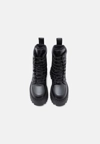 Ζεύγος μαύρων υψηλών χειμερινών μποτών με χοντρές σόλες, κορδόνια και επενδεδυμένα εσωτερικά, που προβάλλεται από πάνω σε λευκό φόντο.