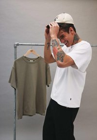 Camiseta blanca de manga corta con cuello redondo, colgando al lado de una camiseta verde oliva en un estante de metal. Se ven pantalones negros.