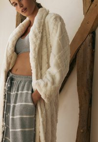 Hvid, kviltet fleecebadekåbe med blød tekstur, udstyret med sjalskrave og brede ærmer. Båret over en grå crop top og stribede sweatpants.