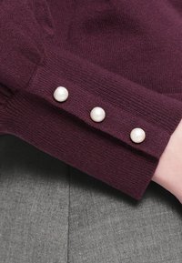 Burgundowy sweter z rękawami ozdobionymi trzema okrągłymi, teksturowanymi guzikami z pereł. Miękka, dziana tkanina z delikatnym prążkowaniem, kontrastująca z szarym tłem.