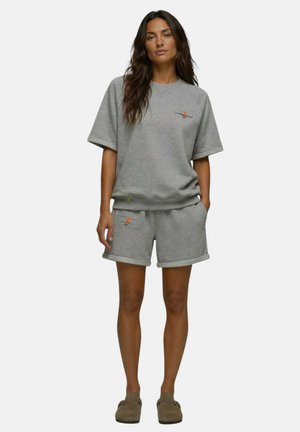 Femme debout portant un haut gris à manches courtes et un short assorti avec de petits détails de fermeture éclair orange et des chaussures marron sans lacets.