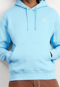 Persona llevando una sudadera Nike de color azul claro con bolsillo frontal y cordones, con las manos metidas en el bolsillo, combinada con pantalones marrones.