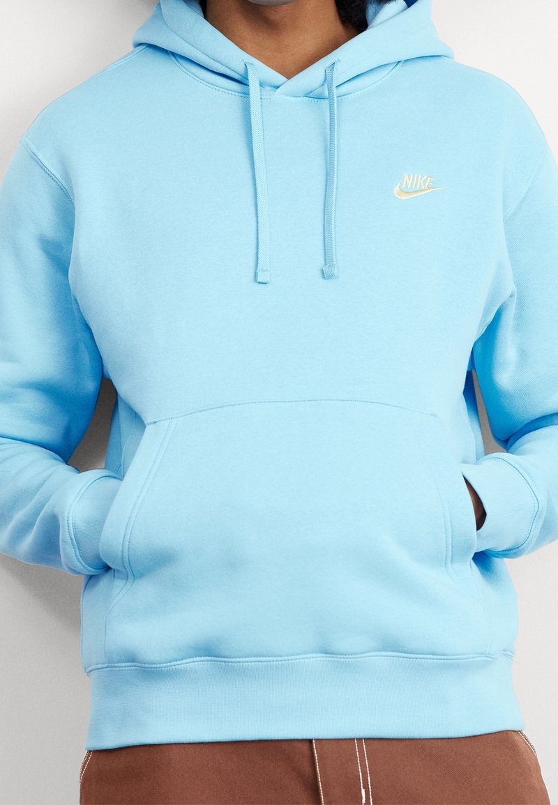 Persona llevando una sudadera Nike de color azul claro con bolsillo frontal y cordones, con las manos metidas en el bolsillo, combinada con pantalones marrones.