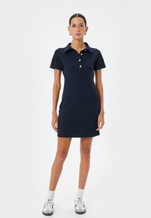 Dunkelblaues Polokleid aus Baumwolle, mit kurzen Ärmeln, einem Kragen und drei goldenen Knöpfen auf der Vorderseite. Glatte Textur und taillierte Form.