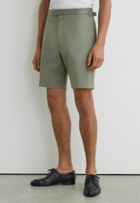 Hombre con pantalones cortos a medida de color oliva claro, camisa blanca por dentro y zapatos negros de piel con cordones, de pie contra una pared blanca y suelo gris.