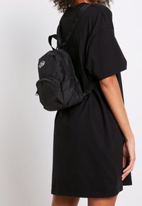 Osoba v černých šatech s krátkým rukávem, která má na zádech malý černý batoh, je zobrazena z boku na jednoduchém pozadí.